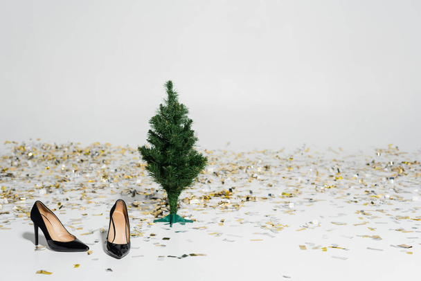 κομψά και μαύρα ψηλοτάκουνα παπούτσια κοντά στο πράσινο χριστουγεννιάτικο δέντρο και αφρώδη κομφετί σε γκρι φόντο - Φωτογραφία, εικόνα