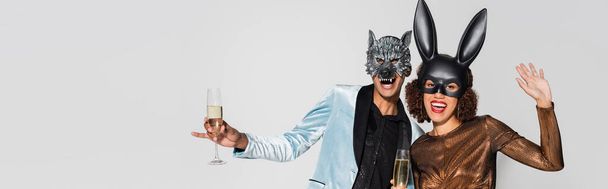 aufgeregtes afrikanisch-amerikanisches Paar in Wolf- und Hasenmasken, Champagner isoliert auf grau, Banner - Foto, Bild