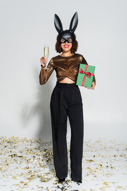элегантная африканская американка в маске кролика с шампанским и рождественским подарком на сером фоне - Фото, изображение
