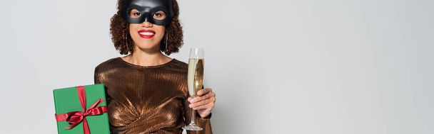 mulher americana africana em blusa elegante e máscara de coelho segurando copo de champanhe e caixa de presente isolado em cinza, banner - Foto, Imagem