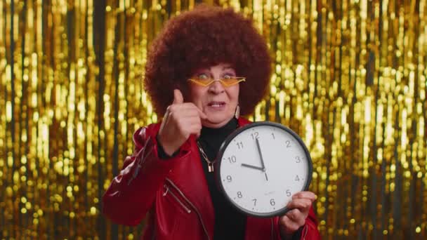 It is your time. Portrait of stylish senior woman showing time on clock watch, ok, thumb up, approve, pointing finger at camera. Elderly grandmother indoors studio shot on shiny disco party background - Footage, Video