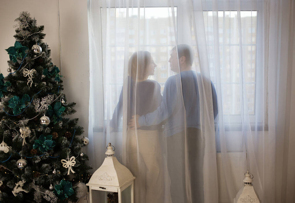 Siluetas de pareja enamorada, hombre mujer, de pie con las espaldas cerca de la ventana detrás de la cortina. árbol de Navidad decorado en la habitación. ambiente de vacaciones, amor, ternura, comodidad. Tema Mañana de Navidad. - Foto, imagen