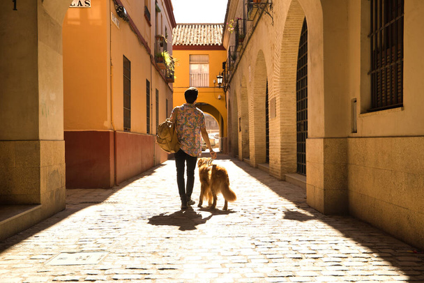 Young Hispanic man on his back with his dog holding the leash walking on a sunny street at sunset. Concept animals, dogs, love, pets, golden. - Foto, Imagen
