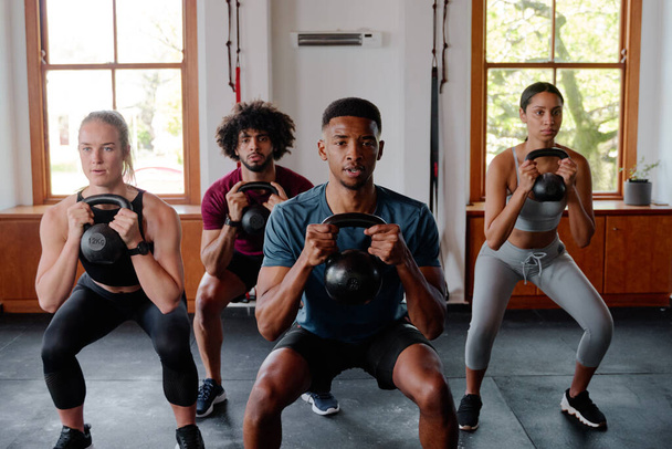 Groupe de jeunes adultes multiraciaux concentrés qui font des squats de kettlebell au gymnase - Photo, image