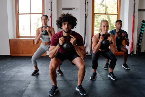 Group of determined multiracial young adults doing kettlebell squats at the gym - Photo, image