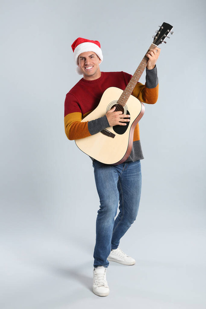 Man in Santa hat playing acoustic guitar on light grey background. Christmas music - Foto, imagen