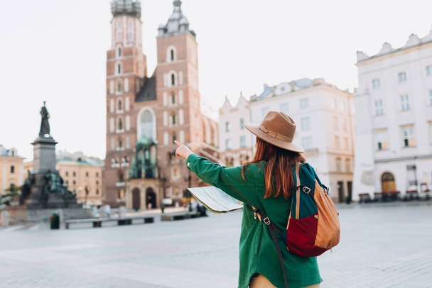 Atrakcyjna turystka poznaje nowe miasto. Ruda kobieta wskazująca palcem i trzymająca papierową mapę na Rynku w Krakowie. Jeździ po Europie jesienią. Bazylika św. Marii - Zdjęcie, obraz