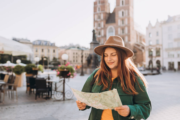 Attractive young female tourist is exploring new city. Redhead girl holding a paper map on Market Square in Krakow. Traveling Europe in autumn. - Foto, imagen