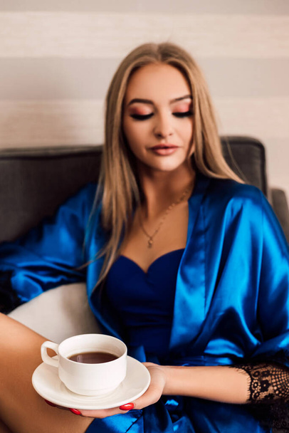 Женщина держит белую чашку кофе и блюдце. Она закрыла глаза и наслаждалась утренним кофе. Синий шелковый халат. Доброе утро. Dawn - Фото, изображение