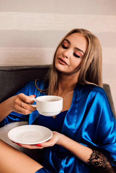 A woman holds a white cup of coffee and a saucer. She closed her eyes and enjoyed her morning coffee. Blue silk robe. Morning. Dawn - Foto, Bild