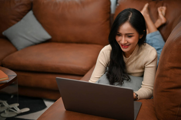 Schöne junge Asiatin, die ihr Wochenende zu Hause verbringt, auf ihrem Sofa liegt, ihren Laptop benutzt, Webblogs liest, Filme anschaut und mit ihren Freunden in den sozialen Medien Kontakte knüpft. - Foto, Bild