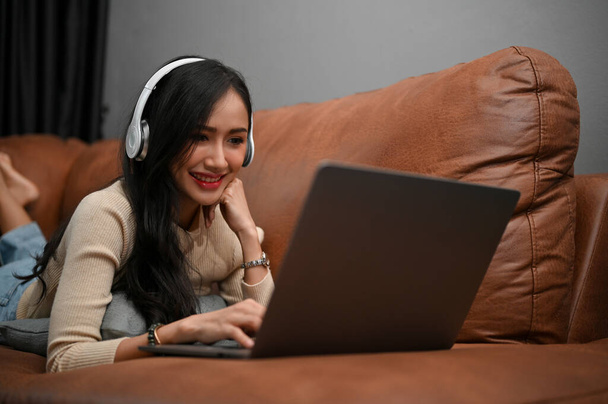 Linda jovem asiática deitada em seu sofá de couro vintage, usando fones de ouvido e assistindo filme em seu laptop. Conceito de hobby e lazer - Foto, Imagem