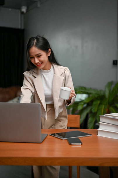 Retrato, mujer de negocios asiática milenaria exitosa y atractiva o gerente ejecutivo de la mujer que comprueba su proyecto de negocio en el ordenador portátil, mirando la pantalla del ordenador portátil, sosteniendo una taza de café. - Foto, imagen