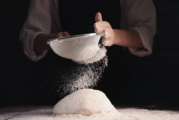 Woman sprinkling flour over dough at wooden table on dark background, closeup - Фото, зображення