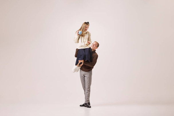 Love, care, dating. Young man in retro style winter fashion clothes holding tender girl on his shoulders isolated on gray background. Christmas, New year, holidays, 2023, party concept - 写真・画像