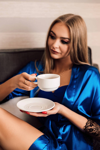 A woman holds a white cup of coffee and a saucer. She closed her eyes and enjoyed her morning coffee. Blue silk robe. Morning. Dawn - Foto, Bild