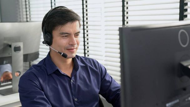 Young competent asian male call center agent working at his computer while simultaneously speaking with customers. Concept of an operator, customer service agent working in the office with headset. - Fotografie, Obrázek