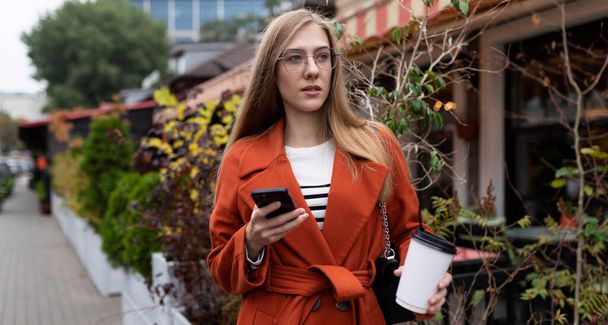 Portakal rengi ceketli genç ve şık bir kadın elinde cep telefonuyla kahve içmek için acele ediyor.. - Fotoğraf, Görsel
