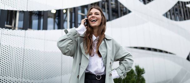 μια νεαρή γυναίκα μιλάει σε ένα κινητό τηλέφωνο με ένα πλατύ χαμόγελο στο πρόσωπό της στο φόντο μιας αλουμινένιας πρόσοψης ενός κτιρίου γραφείων. - Φωτογραφία, εικόνα