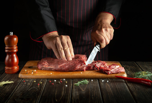 Professional chef cuts raw fresh beef meat on a cutting board before baking or grilling. Working environment in the kitchen of a restaurant or hotel - Foto, afbeelding
