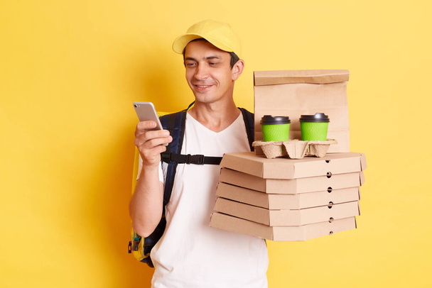 Fast service and best food delivery. Smiling Caucasian delivery man wearing cap and white t shirt holding pizza boxes and takeaway coffee and using mobile phone, isolated on yellow background. - 写真・画像