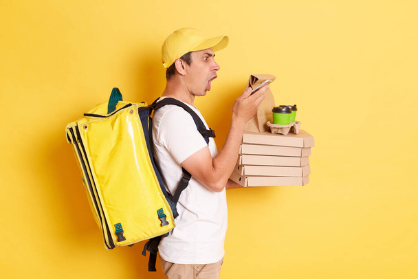 Side view of surprised male courier wearing cap and holding thermo bag holding pizza boxes from restaurant and using smartphone, looking at display with shocked face, isolated on yellow background. - Fotoğraf, Görsel