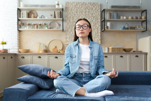 Beautiful Asian woman relaxing at home sitting on sofa and meditating in lotus pose, doing breathing exercises in living room. - Foto, immagini