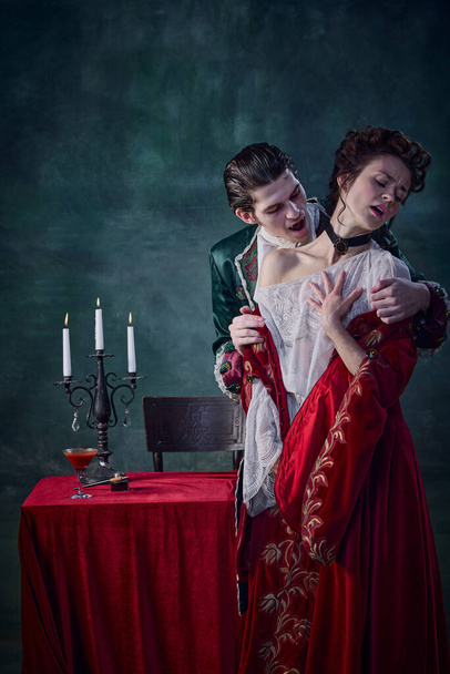 Portrait of young man in image of medieval prince, vampire biting innocent young woman on dark green background. Blood drinking. Concept of Halloween, mystery, gothic art, fantasy, surreal characters - 写真・画像