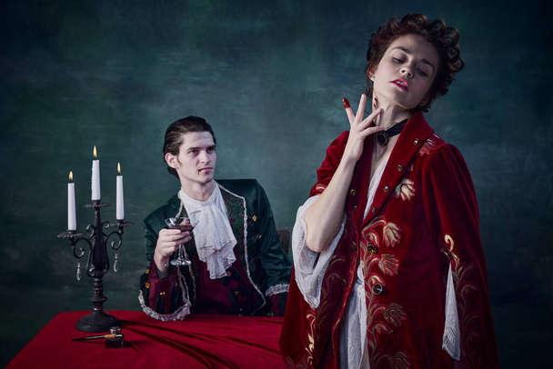 Portrait of fearful family of noble vampires, man and woman in medieval costumes drinking blood over dark green background. Concept of Halloween, mystery, gothic art, fantasy, surreal characters - Photo, image