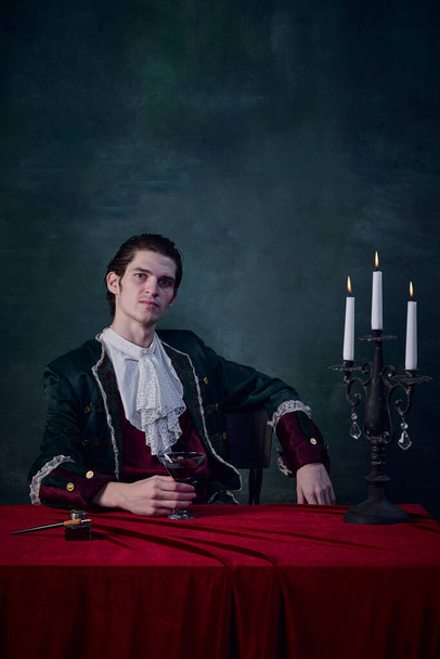 Portrait of nobleman in vintage medieval costume holding glass of red liquid over dark green background. Vampire life. Concept of Halloween, mystery, gothic art, fantasy, surreal characters - 写真・画像