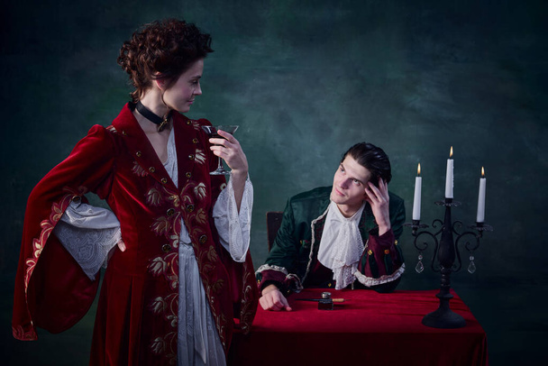 Portrait of immortal lady, woman in image of vampire rising glass of blood near young man, page-boy over dark green background. Concept of Halloween, mystery, gothic art, fantasy, surreal characters - Fotografie, Obrázek
