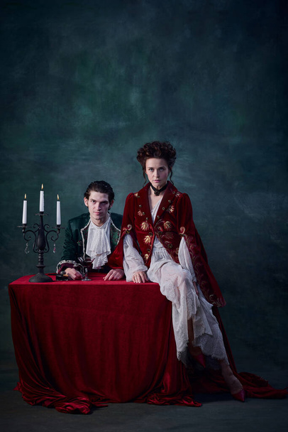 Portrait of mysterious woman and man in medieval costumes sitting over dark green background. Vampire family. Twilight. Concept of Halloween, mystery, gothic art, fantasy, surreal characters - Photo, Image