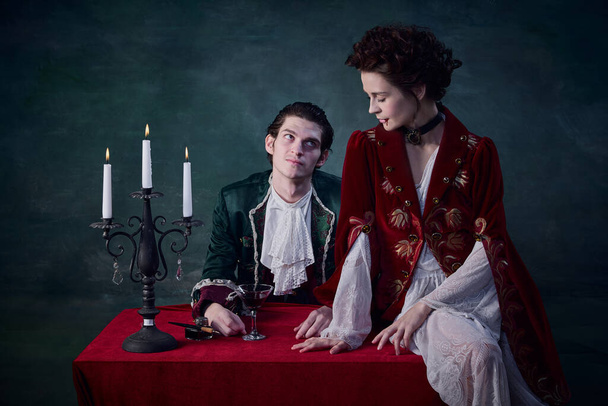 Portrait of elegant woman and man in image of medieval vampires having lunch together over dark green background. Glass of blood. Concept of Halloween, mystery, gothic art, fantasy, surreal characters - Photo, image