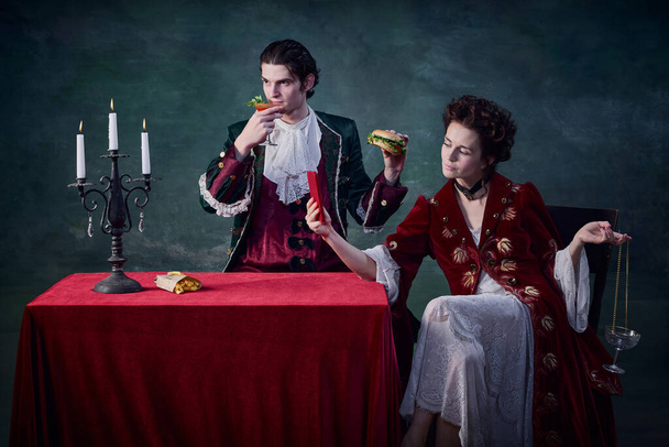 Portrait of man and woman in image of vampires over dark green background. Man eating burger, woman drinking and taking selfie. Combination of eras. Concept of Halloween, fantasy, surreal characters - Fotoğraf, Görsel