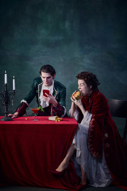 Portrait of people in image of medieval vampires on dark green background. Man looking on phone, woman eating burger. Combination of eras. Concept of Halloween, gothic art, fantasy, surreal characters - Фото, изображение