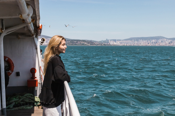 πλαϊνή άποψη του ευτυχισμένη γυναίκα σε μαύρο πουλόβερ κοιτάζοντας τη θάλασσα από πλοίο διέλευσης bosporus στην Κωνσταντινούπολη  - Φωτογραφία, εικόνα