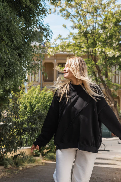 young blonde woman in trendy outfit smiling while walking on street in turkey  - Foto, afbeelding