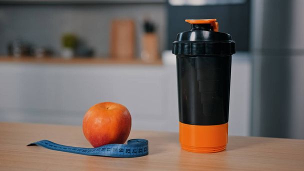 Red apple and tape measure on wooden table at home kitchen plastic bottle with water juice calcium protein food supplements vitamins sport nutrition man athlete takes shaker goes to gym for training - Photo, Image
