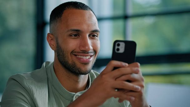 アフリカ系アメリカ人の髭の男が屋内でスマートフォンを使用して男性の雇用主起業家ビジネスマン携帯電話でオフィスに座っている携帯電話のアプリサービスとチャットを入力カメラにネットの笑顔を閲覧 - 写真・画像