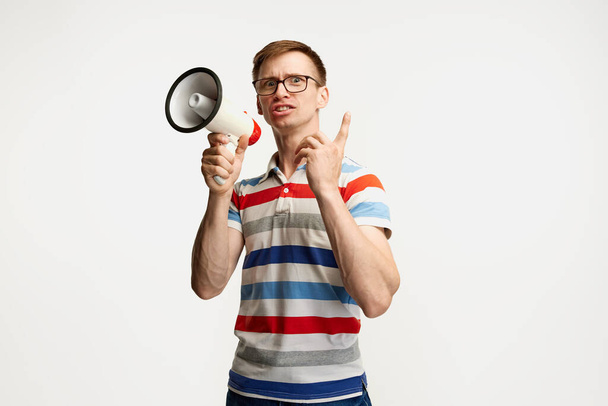 Ritratto di giovane uomo in occhiali in posa con megafono isolato su sfondo bianco da studio. Notizie importanti. Concetto di emozioni, espressione facciale, stile di vita, gioventù. Copia spazio per l'annuncio - Foto, immagini