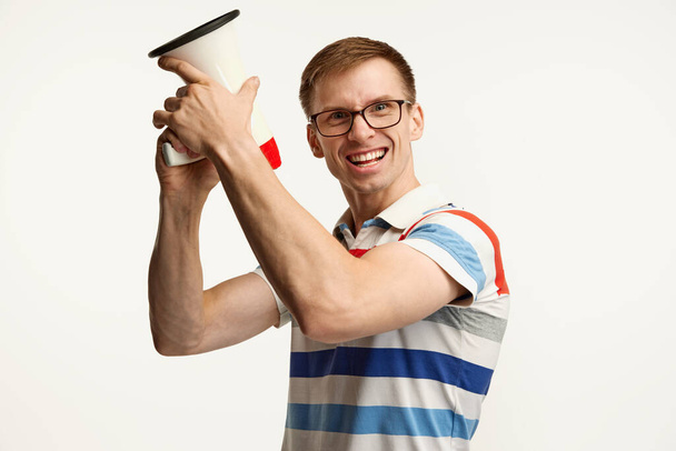 Portrait of young man in glasses posing with megaphone isolated over white studio background. Excitement . Concept of emotions, facial expression, lifestyle, youth. Copy space for ad - Фото, зображення