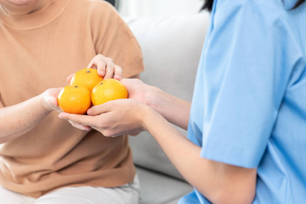 Молодой сиделка раздает апельсины своему довольному старшему пациенту в гостиной. Услуги по уходу за престарелыми на дому. - Фото, изображение