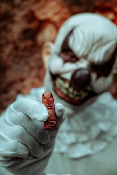 detailní záběr strašidelného zlého klauna, oblečeného v šedém kostýmu s bílým rukávem, ukazujícího zakrvácený amputovaný prst, před starou rezavou vnější zdí opuštěného domu za soumraku - Fotografie, Obrázek
