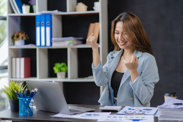 A young businesswoman excited about her business success raises both her arms as she sits at a desk in a small office. - Фото, изображение