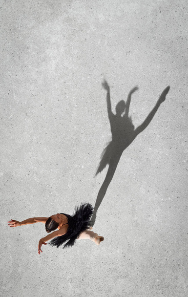 Porträt einer jungen Ballerina im schwarzen Kleid, die isoliert vor grauem Hintergrund auftritt. Ansicht von oben. Schattenelement. Höhenflug. Konzept des klassischen Balletts, Inspiration, Schönheit, Tanz, Kreativität - Foto, Bild