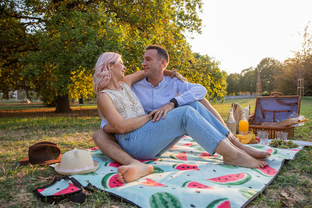 Обнятая пара, сидящая на одеяле, намеренная на веганском пикнике, смотрит друг другу в глаза с любовью. - Фото, изображение