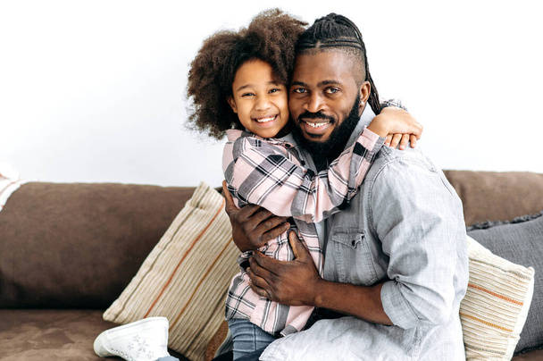 Portrait d'un heureux père afro-américain et sa petite fille mignonne aux cheveux bouclés préscolaires se serrent dans leurs bras alors qu'ils sont assis sur un canapé dans le salon, passent du temps ensemble, sourient à la caméra - Photo, image