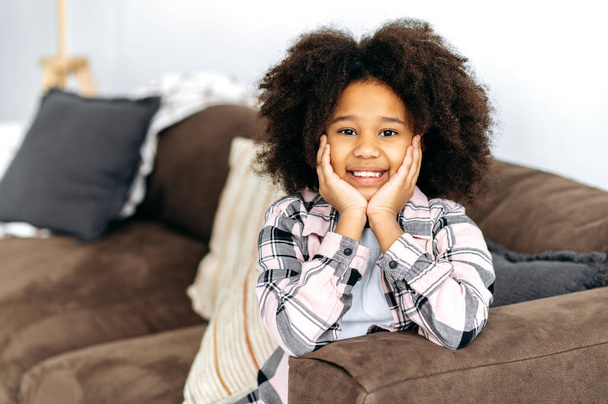 Портрет веселой милой африканской маленькой девочки дошкольного возраста с вьющимися волосами, в повседневной одежде, сидит на диване в гостиной, позирует перед камерой, счастливо улыбаясь - Фото, изображение
