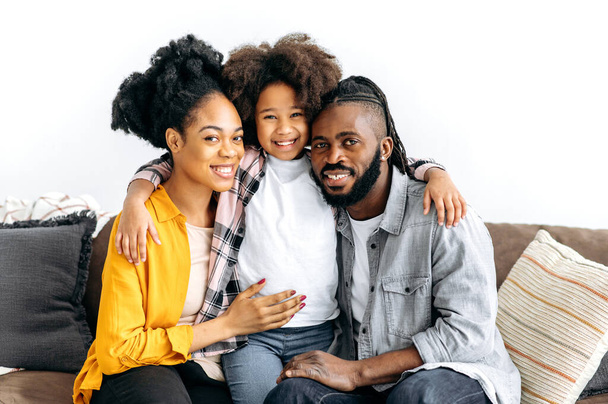 Happy family portrait. Amazing african american family, father, mother and little daughter hugging while sitting on a couch in living room and smiling into camera, spending time together at home - Photo, Image