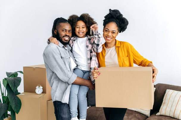Új lakás, áthelyezés. Boldog afro-amerikai család, apa anya és aranyos lányuk, beköltöztek az új lakásukba, dobozokkal a kezükben, a lány az apák karjában kulcsokat mutat, mosolyognak a kamerába - Fotó, kép
